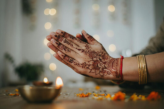 Close up of hands adorned with traditional intricate mehndi henna designs pressing together in a cultural gesture of prayer or greeting near flickering oil lamps and scattered vibrant flower petals