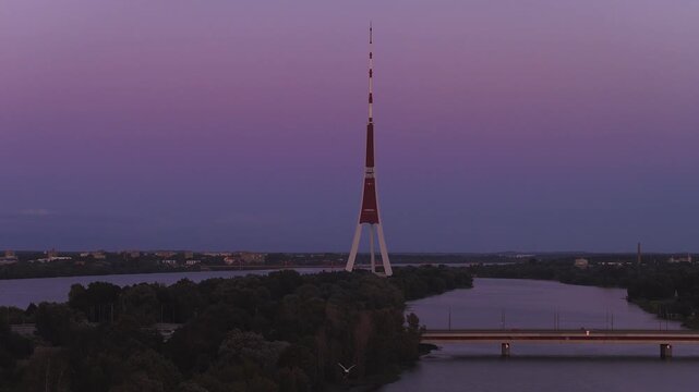 Aerial dusk view of Riga TV Tower on Zakusala in the Daugava River. A low bridge carries cars, headlights move, water reflects, sky shows purple pink gradient.
