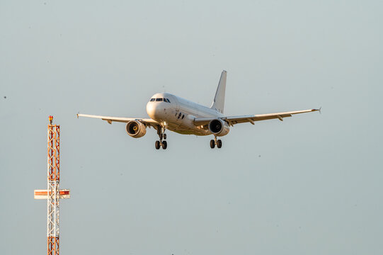 A passenger jet flies on approach past a red and white navigation mast at Brussels Airport in Belgium in soft evening light.