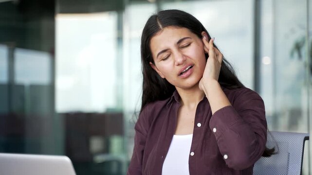 Young businesswoman suffering from ear pain while working on laptop at workplace in modern office. Female employee touches her ear feels discomfort, inflammation, symptoms of ear infection. Close up.