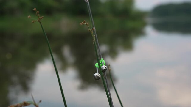 Close-up 4K footage of a small fishing bite alarm bell attached to the tip of a fishing rod beside a pond. The bell is used as a simple bite indicator to alert an angler when a fish pulls the line.
