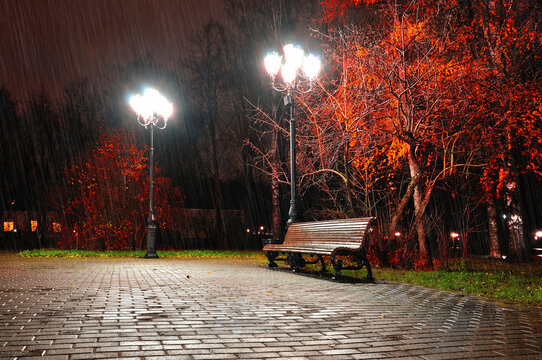 Autumn night landscape of deserted park - a wooden lonely bench lit by street lamps under falling rain in the evening