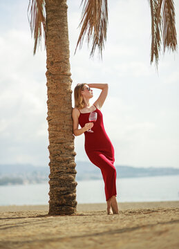 Elegant young woman in red dress, holding glass of wine, having fun and enjoying her vacation on seashore with the city in background