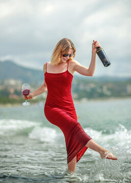 Elegant young woman in red dress, holding glass of wine, having fun and enjoying her vacation on seashore with the city in background