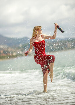 Elegant young woman in red dress, holding glass of wine, having fun and enjoying her vacation on seashore with the city in background