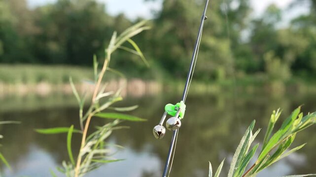 Close-up 4K footage of a fisherman attaching a small metal bite alarm bell to the tip of a fishing rod beside a pond. The bell is used as a simple fishing bite indicator to alert the angler fishing
