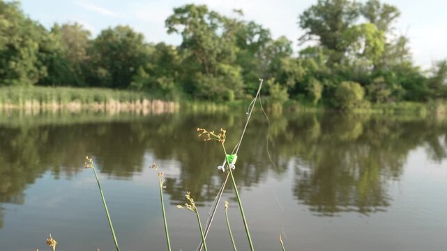 Close-up 4K footage of a fisherman attaching a small fishing bite alarm bell to a rod beside a calm pond. The angler tests the setup by shaking the fishing rod, showing a simple bell indicator signal
