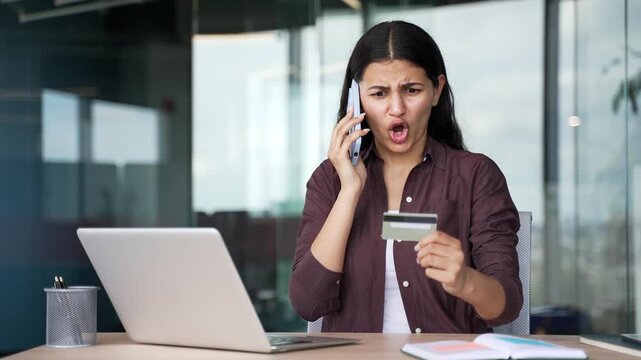 Frustrated young businesswoman arguing on mobile phone while holding credit card in business office. Angry female customer dealing with banking issue, declined payment or unexpected transaction error.