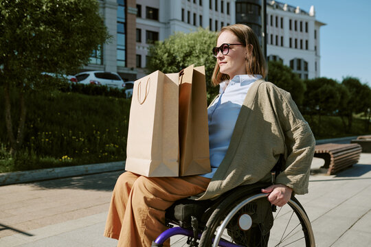Caucasian young adult woman with disability carrying shopping bags while moving in wheelchair outdoors, representing accessible retail and independent living. For inclusion marketing