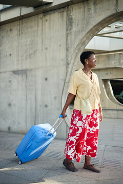 Vertical shot of Black middle aged woman pulling wheeled suitcase while waiting outdoors, representing solo travel and mobility. Ideal for tourism ads, vacation booking, travel insurance