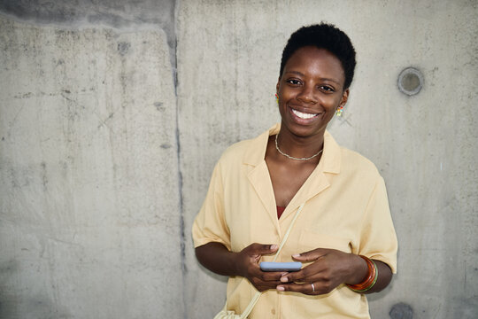 Portrait of young adult Black woman holding smartphone and smiling at camera against concrete wall, conveying mobile communication and confidence. Useful for digital lifestyle marketing