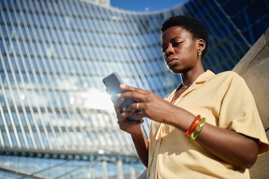 Low angle shot of young Black woman using smartphone outside office building, managing mobile communication and digital workflow. Ideal for business technology, networking, fintech marketing