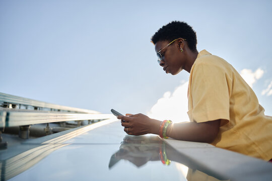 Low angle shot of young Black woman using smartphone beside solar panels, managing clean energy workflow and mobile monitoring. Ideal for renewable technology, sustainability marketing