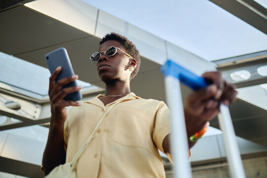 Low angle shot of young Black woman checking smartphone while pulling wheeled suitcase during transit, representing mobile travel planning. Ideal for tourism apps, airport services, digital booking