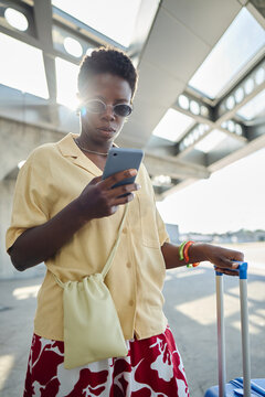 Vertical shot of young Black woman checking smartphone while traveling with rolling suitcase at transit terminal. Ideal for mobile travel app, tourism, commute, connectivity marketing