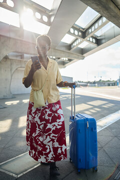 Vertical shot of young Black woman checking smartphone while pulling suitcase at transit station, representing mobile travel planning. Ideal for tourism ads, booking apps, commuting