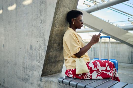 Wide shot of young Black woman sitting with suitcase and using smartphone while waiting at transit terminal, representing mobile travel planning. Ideal for tourism apps or booking ads