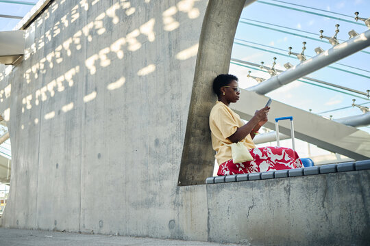 Wide shot of young Black woman checking smartphone beside rolling suitcase at transit terminal, waiting for trip updates. Ideal for travel app, mobility service, tourism campaign use
