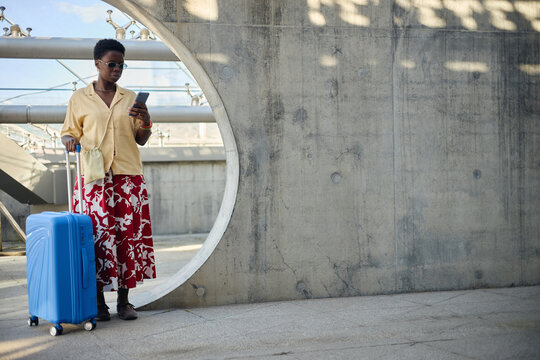 Full length shot of young adult Black woman checking smartphone while standing with rolling suitcase in transit space. Ideal for travel booking, mobile app, tourism campaign use