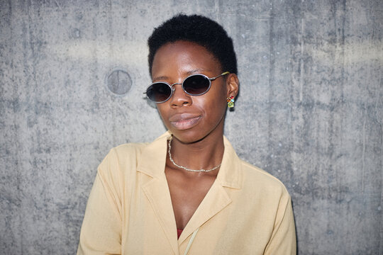 Portrait of young adult Black woman wearing sunglasses and facing camera against concrete wall, conveying confident personal branding. Ideal for fashion marketing, identity campaigns