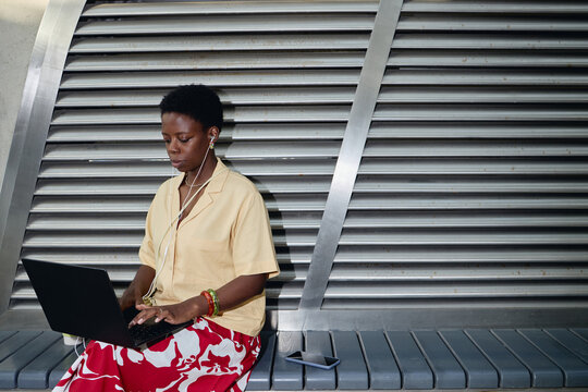 Medium shot of young Black woman working on laptop while listening through earphones in urban public space, representing mobile productivity. Ideal for remote work or digital nomad marketing