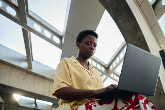 Low angle shot of young Black woman working on laptop in public building, managing remote tasks and digital communication. Ideal for hybrid work, productivity, mobility marketing