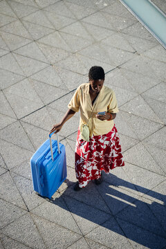 Vertical shot of Black young adult woman pulling suitcase while checking smartphone on paved walkway, suggesting solo travel planning. Ideal for tourism apps, mobility services, trip booking