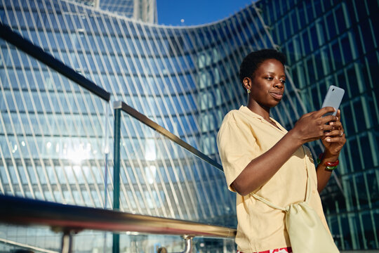Young African American woman using smartphone outside modern office building, checking mobile communication and digital services. Low angle shot, ideal for fintech, networking, urban tech marketing