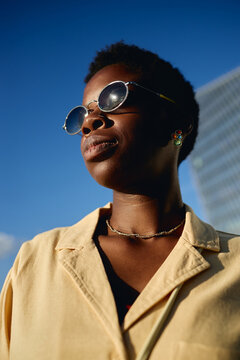Vertical shot of Black young adult woman wearing sunglasses, facing sunlight in city, conveying confidence and modern urban lifestyle. Ideal for fashion branding, identity campaigns