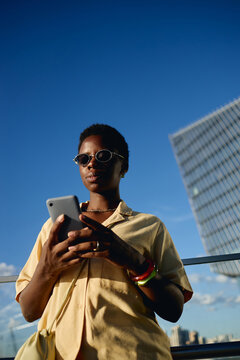 Vertical low angle shot of young Black woman using smartphone outdoors near office tower, managing mobile communication and urban workflow. Ideal for digital lifestyle or business app marketing