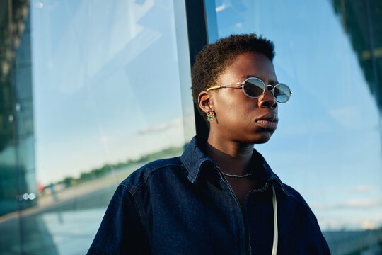 Close up of young adult Black woman wearing sunglasses and standing outdoors near glass facade, conveying urban confidence and modern identity. Ideal for fashion branding and lifestyle campaigns