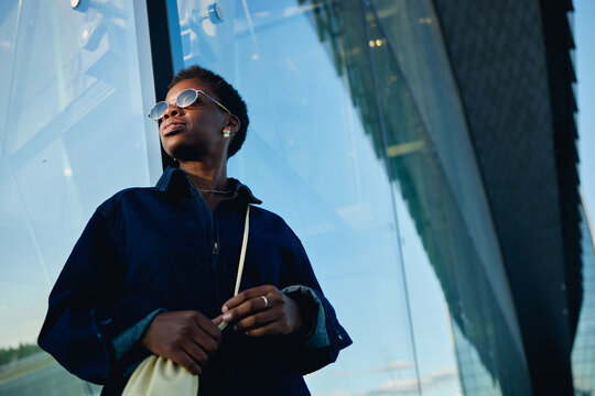 Low angle shot of young Black woman standing by glass office building, looking away with confident expression, representing leadership and urban career branding. For business identity campaigns