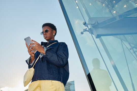 Young adult Black woman using smartphone beside modern office building, managing mobile communication and digital workflow. Ideal for business technology, connectivity, urban lifestyle marketing