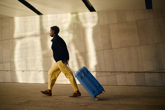 Full length shot of Black young adult woman pulling wheeled suitcase while walking through transit terminal, representing business travel and mobility. Ideal for tourism marketing