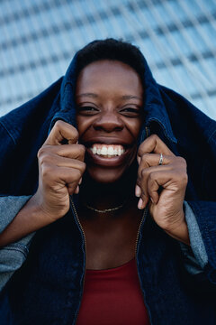 Vertical portrait of young Black woman smiling at camera while holding jacket over head, expressing confidence and playful individuality. Useful for fashion marketing, lifestyle branding
