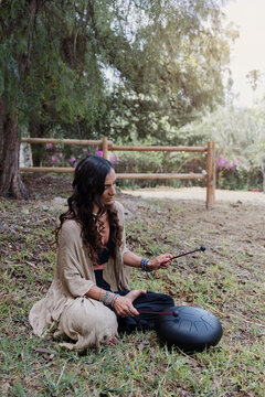 young latin Woman playing a steel tongue drum while seated on grass wearing colorful and practicing sound therapy for relaxation and meditation outdoors in Latin America