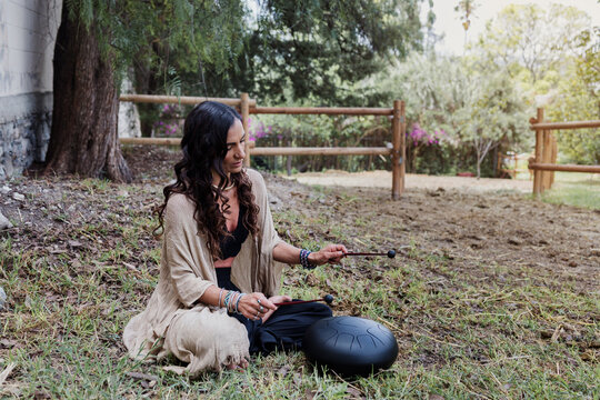young latin Woman playing a steel tongue drum while seated on grass wearing colorful and practicing sound therapy for relaxation and meditation outdoors in Latin America