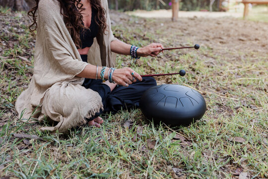 young latin Woman playing a steel tongue drum while seated on grass wearing colorful and practicing sound therapy for relaxation and meditation outdoors in Latin America