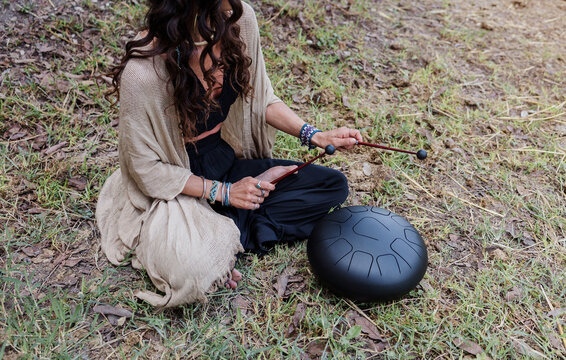 young latin Woman playing a steel tongue drum while seated on grass wearing colorful and practicing sound therapy for relaxation and meditation outdoors in Latin America