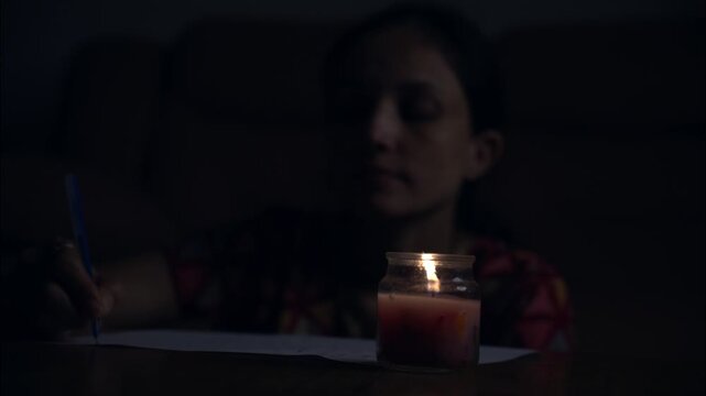 A moody low-light footage of a woman writing on paper beside a burning candle in a quiet room.