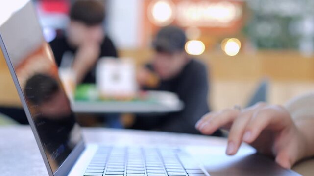 Hands type on laptop in bright cafe. Background shows blurred people. Focus on keyboard and fingers. Suitable for remote work or digital lifestyle themes