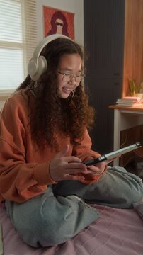 Vertical video of Asian teenager wearing headphones and glasses, sitting on bed and using tablet. Footage is ideal for promoting educational apps, online tutoring, and digital learning