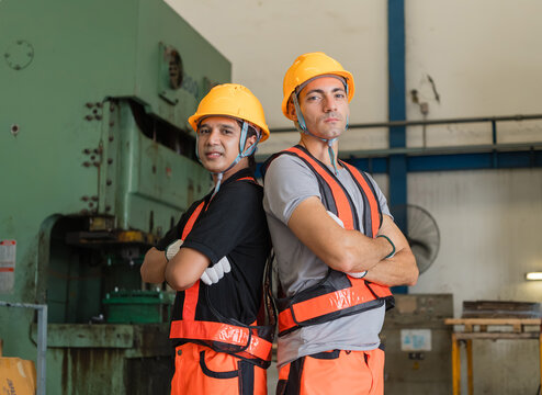 Two workers stand back to back, arms crossed, beneath roof trusses and a green power press inside a tall metalworking factory