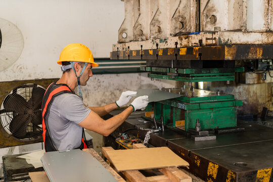 Machine operator in yellow hard hat and vest guides a metal sheet into a punch press die on the factory floor