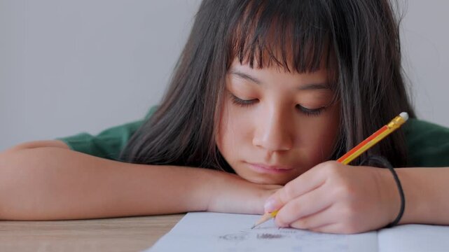 Close up selective focus of demotivated Asian kid girl resting her head on a desk in daylight. She looks overburdened by too much homework, illustrating academic pressure and childhood study burnout.