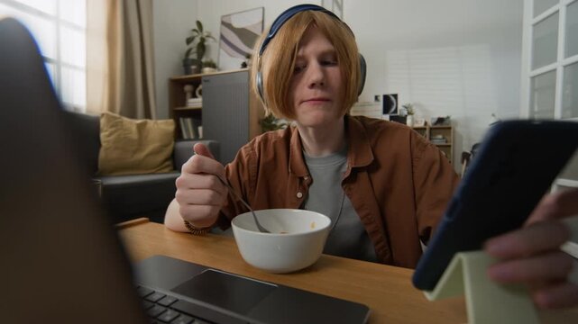 Caucasian teenager wearing headphones eating cereal, using smartphone and typing on laptop at home. Perfect for marketing campaigns targeting Gen Z digital habits and remote learning