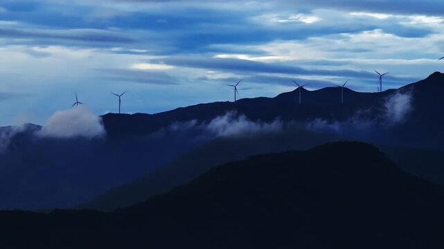 Wind turbines stand on mountain ridges surrounded by clouds during twilight, representing renewable clean energy.