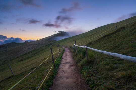 Hiking trail path with wooden fence on green mountain slope at sunset in Dolomites, Italy. Alpine dirt footpath leading to misty hill top, landscape with high altitude grass field at night. Summer