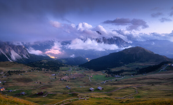 Alpine valley view with village lights and wooden huts at dusk in Dolomites, Italy. High altitude mountain landscape with low clouds covering rocky peaks at night. Twilight over green hills in summer