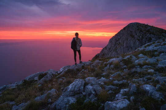 Woman hiker with backpack standing on rocky mountain top at colorful sunset in Croatia. Female tourist looking at vibrant red and peak sky and sea view from mountain peak, outdoor activity in summer